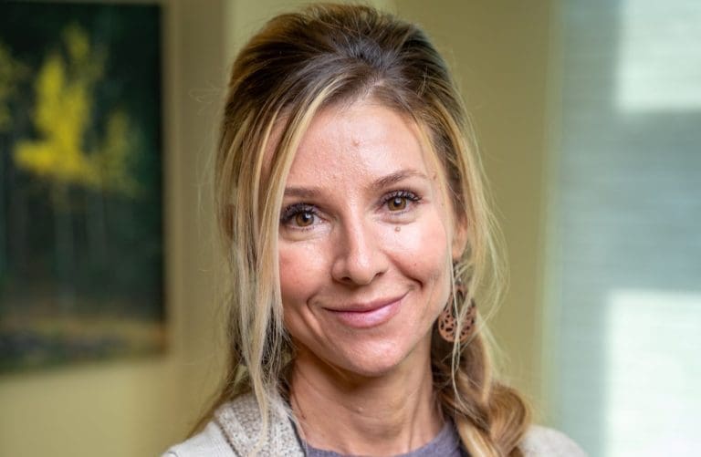 A woman with long blonde hair in a loose braid smiles at the camera. She is wearing brown earrings and a beige top. The softly blurred background hints at a window and a painting of trees, reflecting her passion for affordable health care.