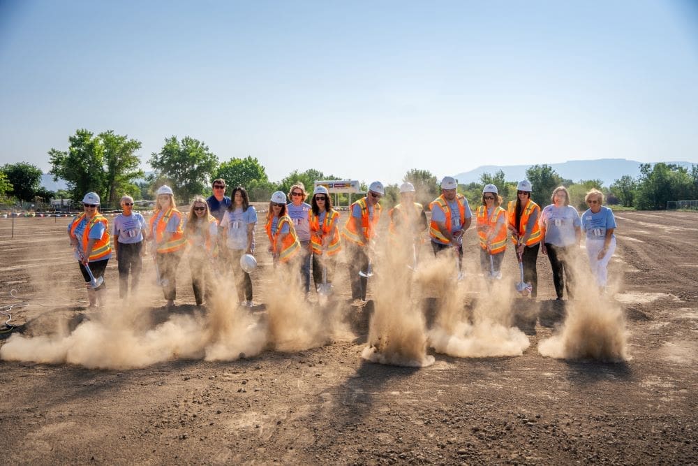 A group of people wearing safety vests and helmets break ground on a Grand Junction Health Care construction site, kicking up clouds of dirt as they shovel. The outdoor setting features a clear blue sky and trees in the background.