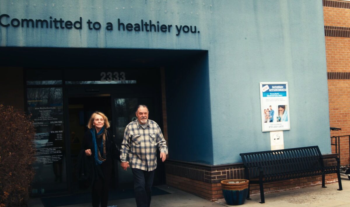 An older woman and man walk out of a building with a sign above the entrance reading, Committed to a healthier you. A bench, poster, and potted plant are next to the entrance of this Grand Junction Health Care facility.