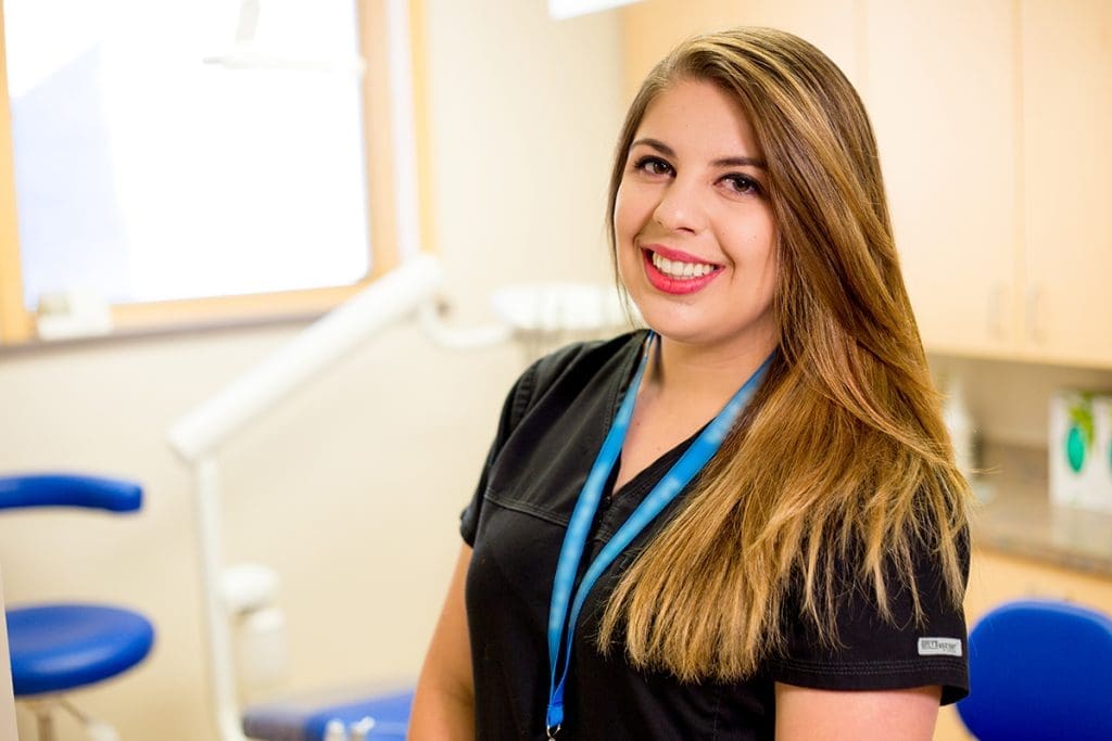 A woman wearing black scrubs and a blue lanyard smiles while standing in a brightly lit Grand Junction Health Care dental office, with dental equipment in the background.