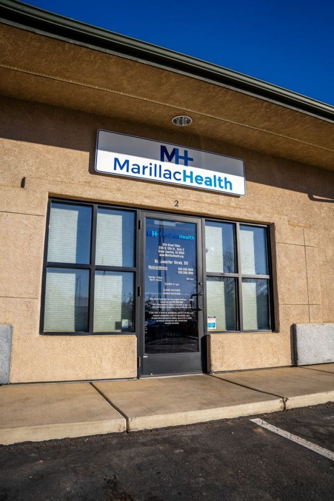 A tan building with large windows and a glass door displays a sign reading MarillacHealth above the entrance, offering affordable health care. The door lists clinic information, and there is a parking spot in front.
