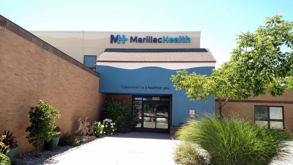 The entrance of MarillacHealth, an affordable health care facility in Grand Junction, features a blue and beige exterior, landscaped plants, and a welcoming sign above the door that reads Committed to a healthier you.
