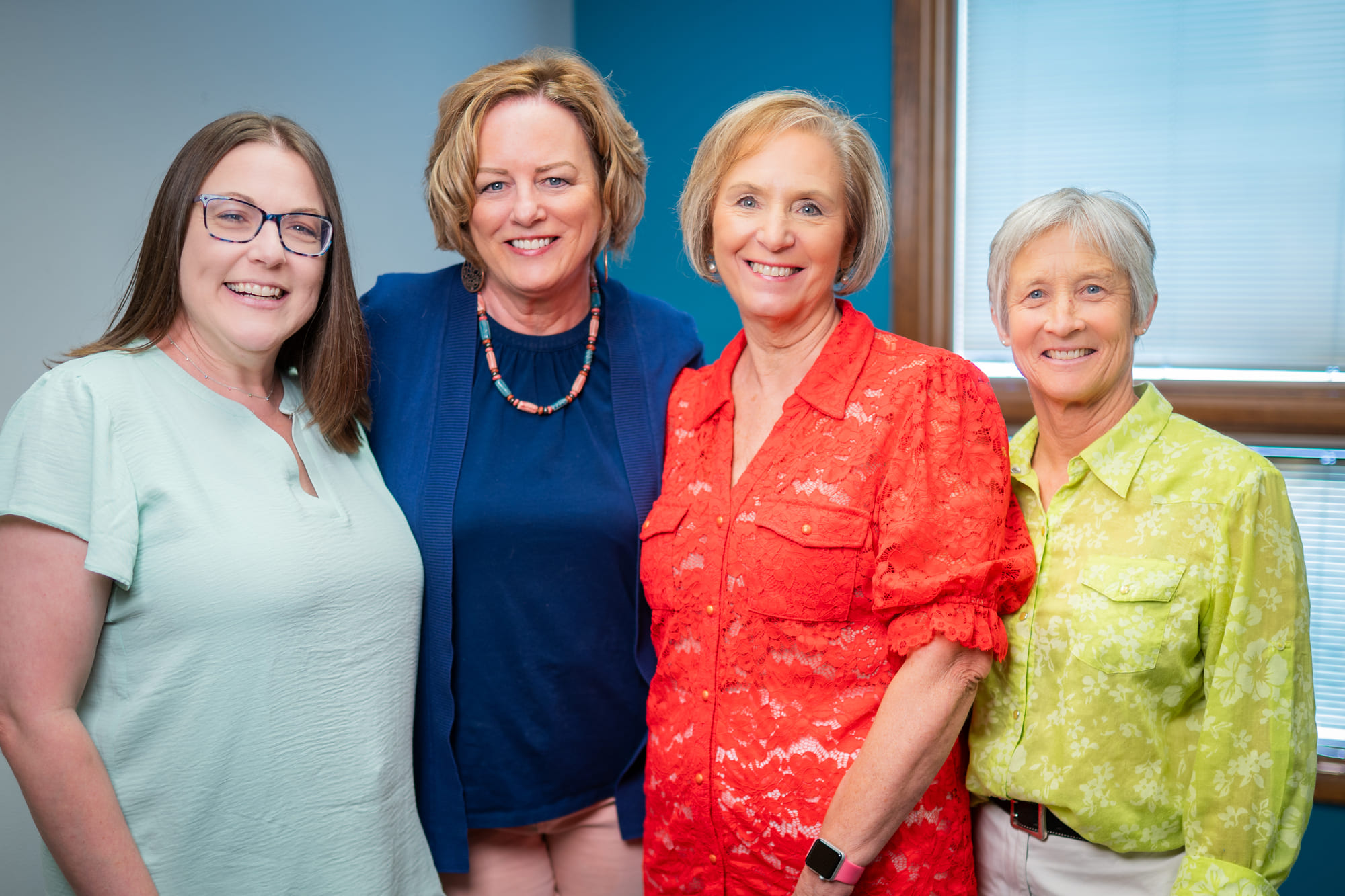 Four smiling women stand together indoors in front of a window, wearing brightly colored blouses. They appear relaxed and happy in a well-lit room with blue walls, reflecting the friendly spirit of Montrose Health Care.