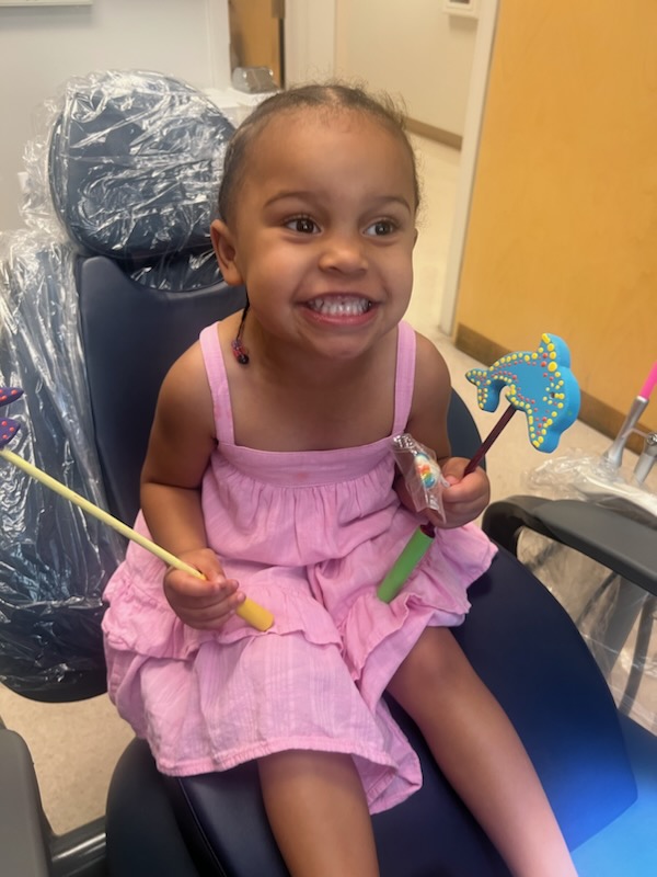 A young girl in a pink dress smiles widely while sitting in a Montrose Health Care dental chair, holding a toy wand in one hand and a blue decorated cookie shaped like a dolphin in the other.