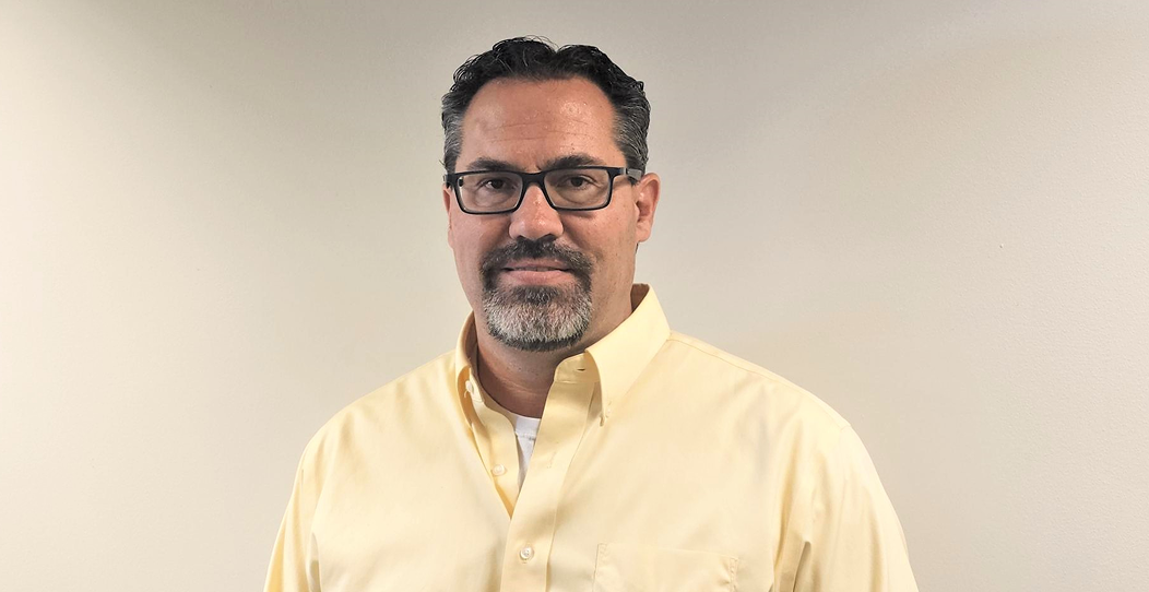 A man with short dark hair, glasses, and a salt-and-pepper goatee, wearing a light yellow button-up shirt, stands in front of a plain off-white wall. He looks at the camera with a neutral expression, reflecting Montrose Health Care’s approachable professionalism.