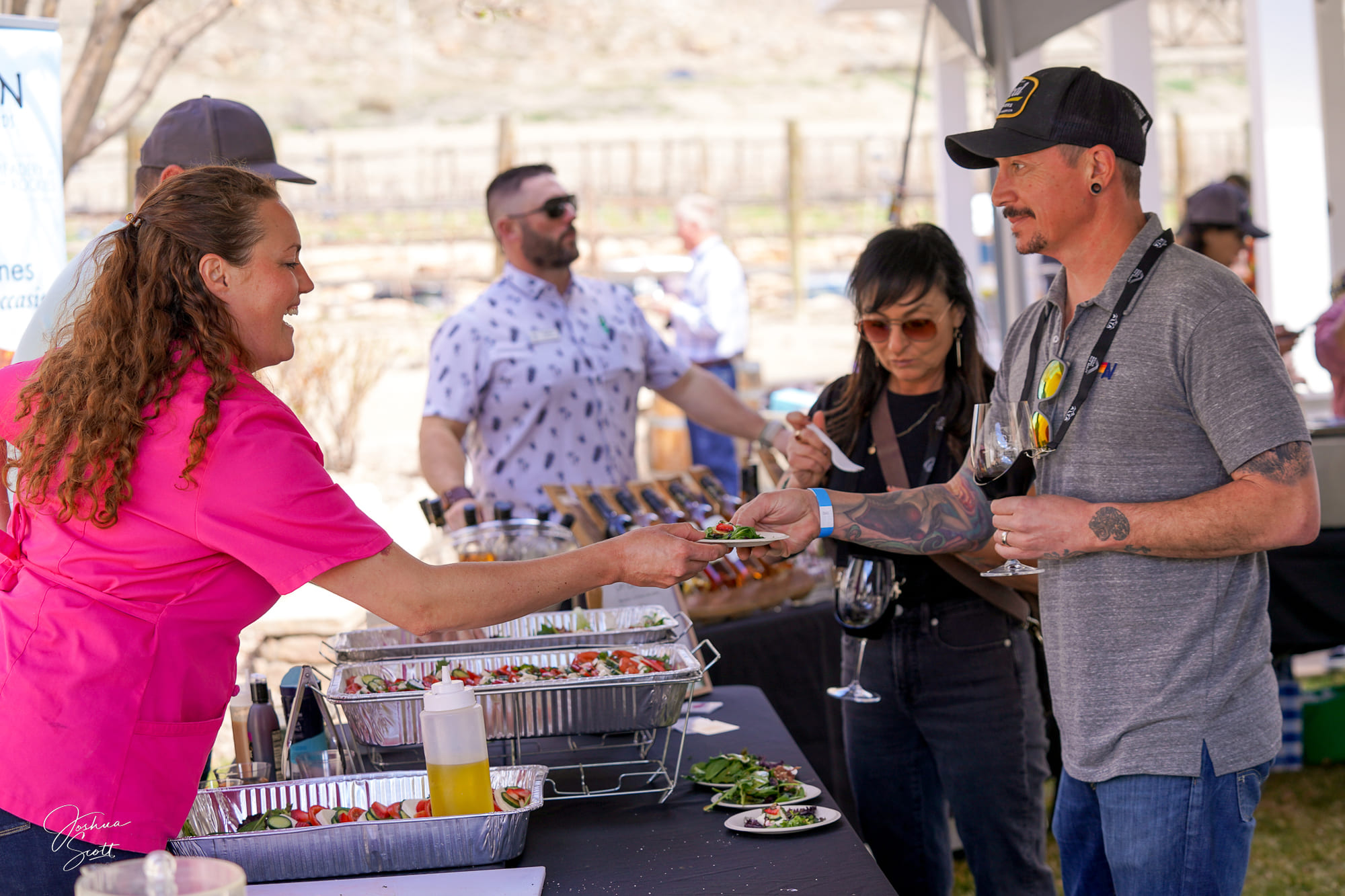 A woman in a pink shirt serves food to a man with tattoos at an outdoor event, hosted by Montrose Health Care, while others stand nearby holding wine glasses. A buffet with trays of food is set up on the table.