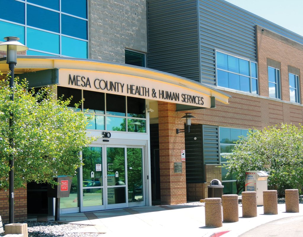 The entrance to the Mesa County Health & Human Services building in Grand Junction, with glass doors, large windows, a curved yellow awning, and surrounding greenery on a sunny day, highlights access to affordable health care in the community.