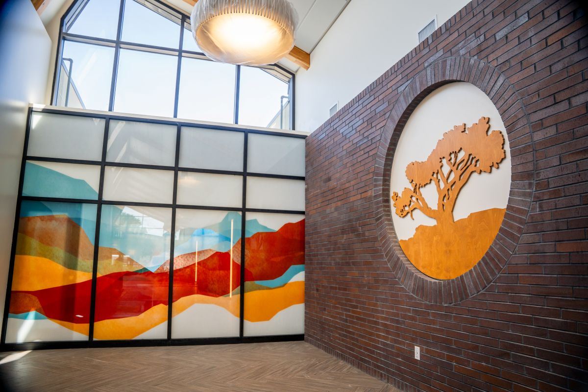 A modern interior features a large window, colorful abstract wall art with layered shapes, and a circular brick accent with a metal tree design—an inviting space that reflects the warmth of Grand Junction Health Care. Natural light fills the space.
