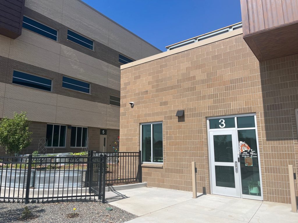 A tan brick school building with a door labeled 3, a security camera, and large windows. A black fence runs along the walkway, and part of another Montrose Health Care building is visible in the background. The sky is clear and blue.