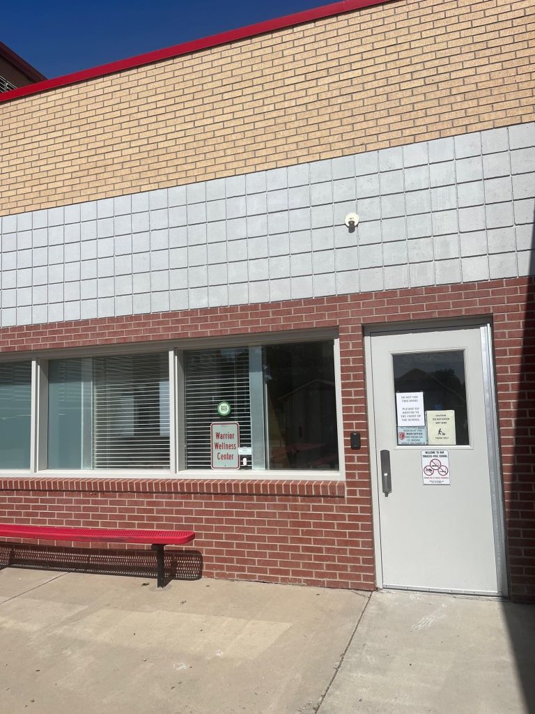 A brick building with a window and a white door labeled Warrior Wellness Center. A red metal bench sits outside, and signs for Affordable Health Care are posted. The building exterior is tan, gray, and red.