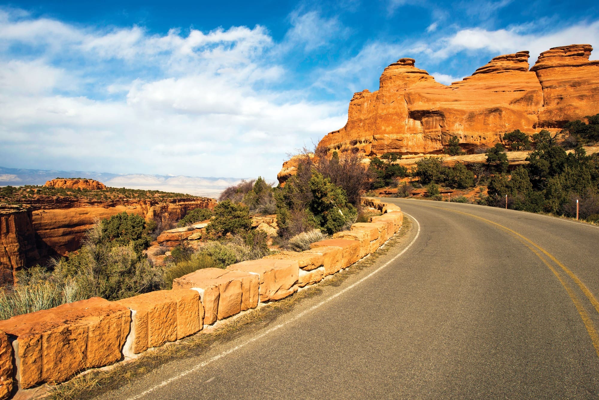 A paved road curves beside orange-red rock formations and cliffs under a bright blue sky with scattered clouds, surrounded by desert vegetation—just miles from Montrose Health Care and quality, affordable health care options nearby.