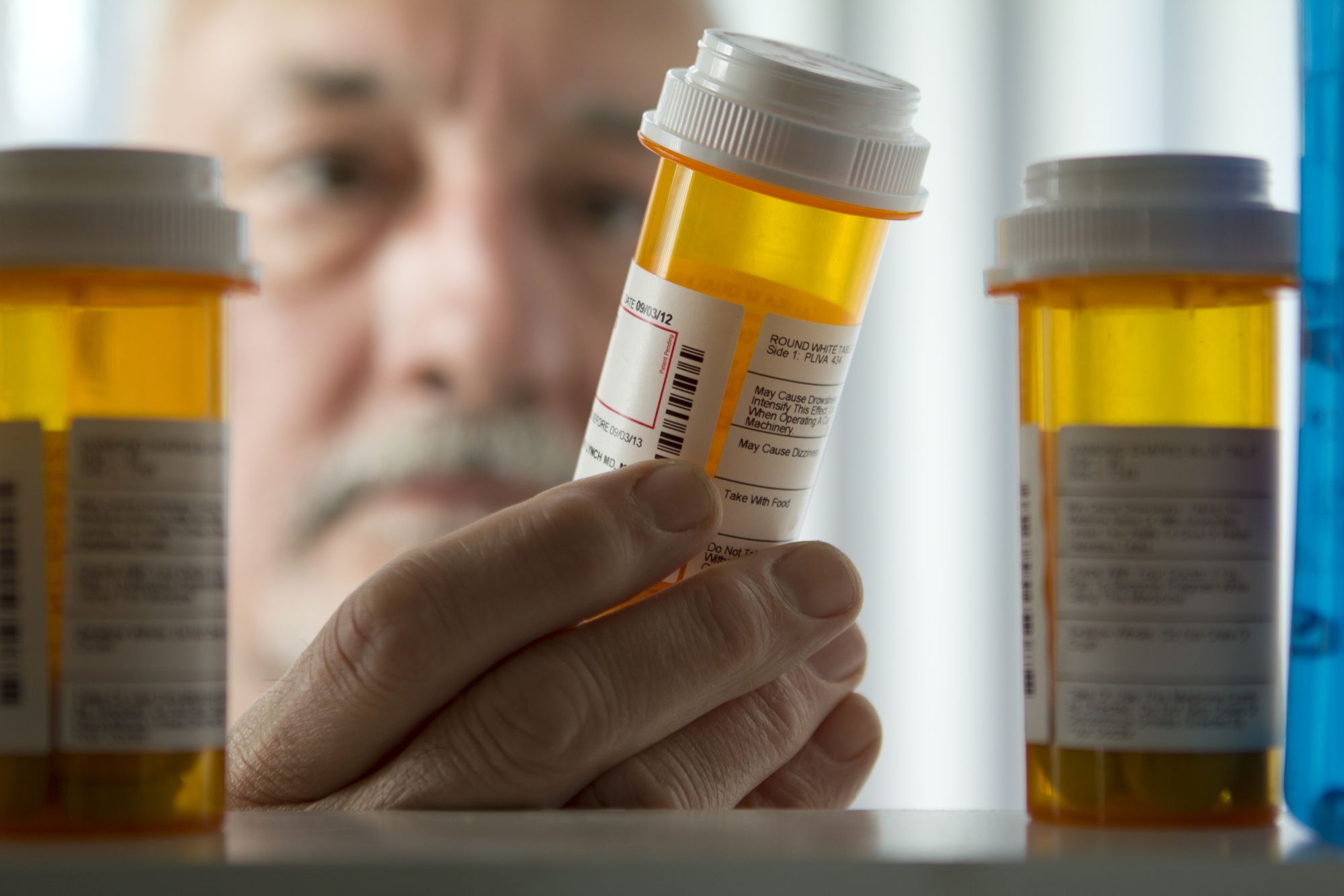 A person’s hand reaches for a prescription pill bottle on a shelf, with more similar bottles nearby; the person’s face is out of focus in the background, highlighting the importance of access to healthcare.