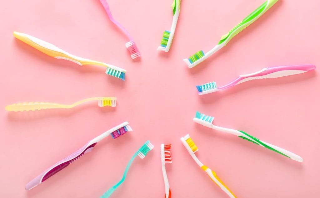 Twelve colorful toothbrushes arranged in a circle on a pink background, their bristles pointing toward the center—a vibrant reminder of good oral health and its importance for overall wellness at Montrose Health Care.