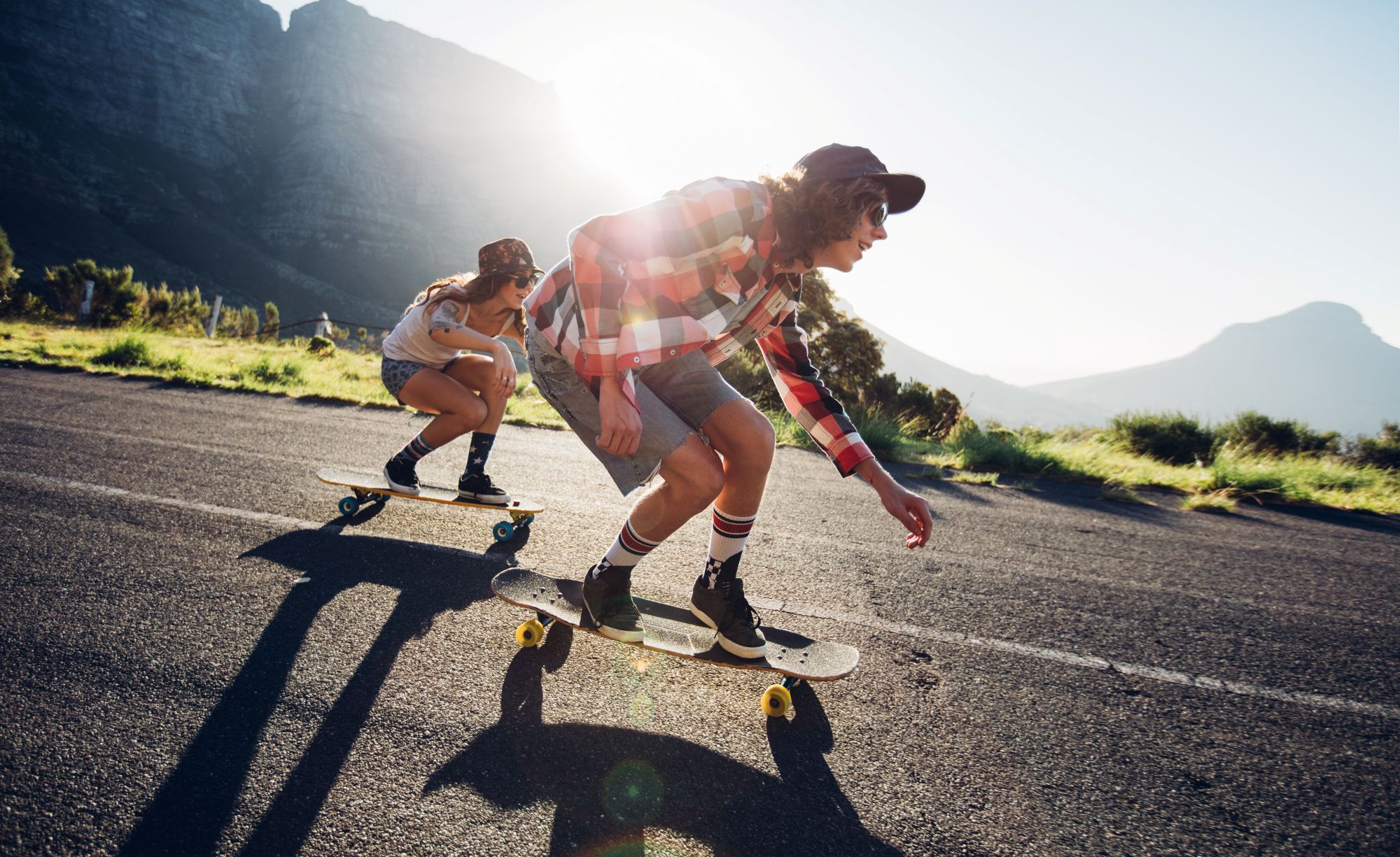 Two people skateboard down a paved road in the sunlight, surrounded by mountains and greenery—a perfect reminder to enjoy the outdoors and stay healthy with affordable health care options from Montrose Health Care.