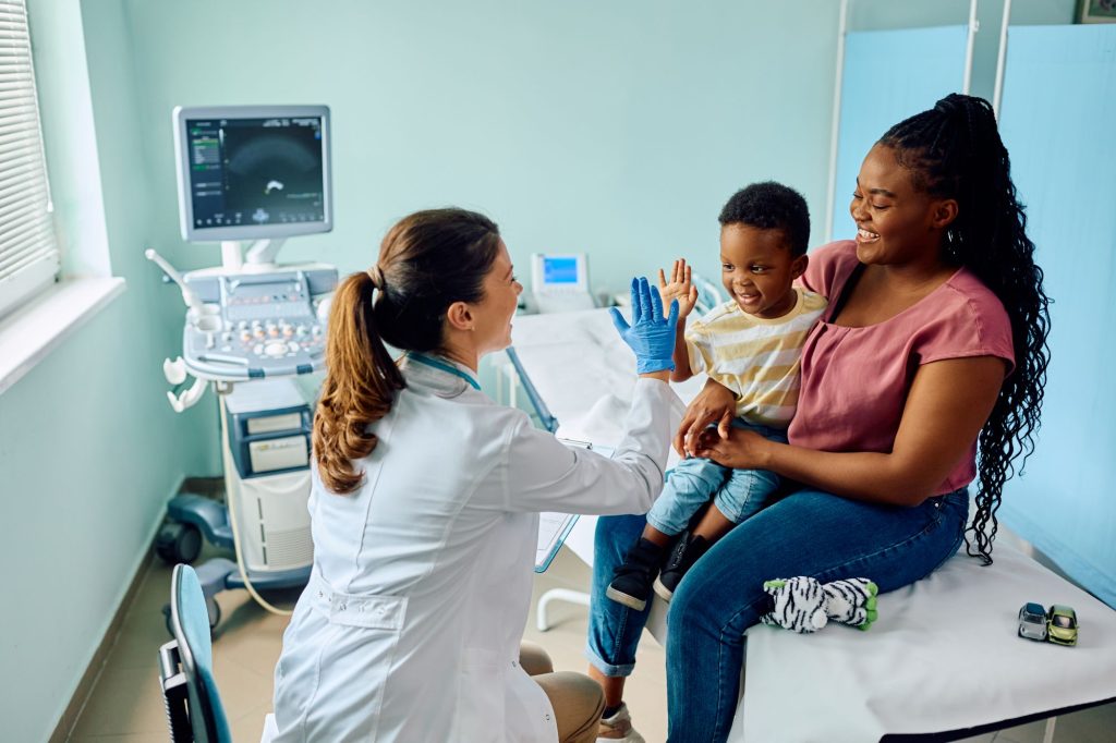 A doctor wearing gloves gives a high-five to a smiling young boy sitting on an exam table with his mother in a Montrose Health Care office; medical equipment is visible in the background.