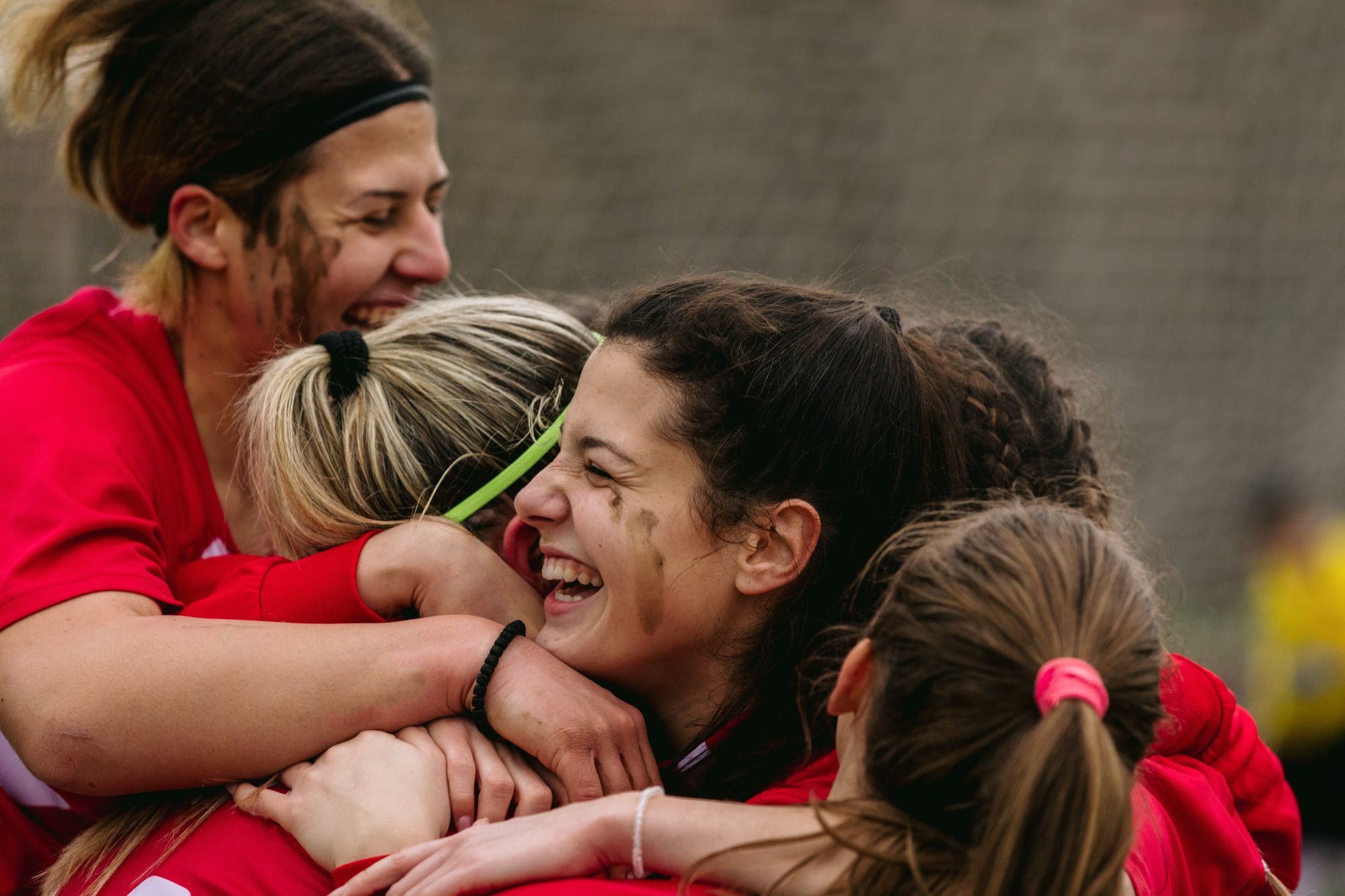 Four women in red uniforms, mud on their faces, joyfully hug and celebrate together on a sports field, smiling and laughing—a true display of teamwork and happiness, just like the spirit found at Grand Junction Health Care.
