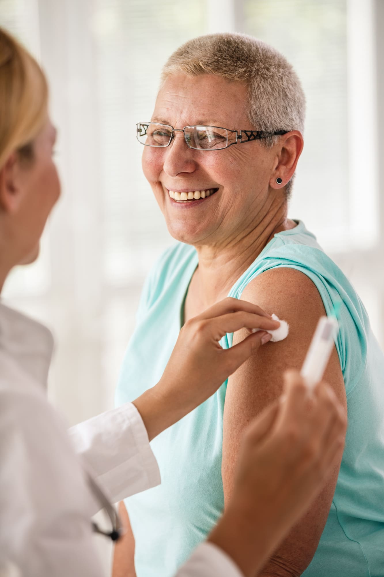 A smiling older woman with short gray hair and glasses receives a vaccine shot in her upper arm from a healthcare professional at Montrose Health Care.