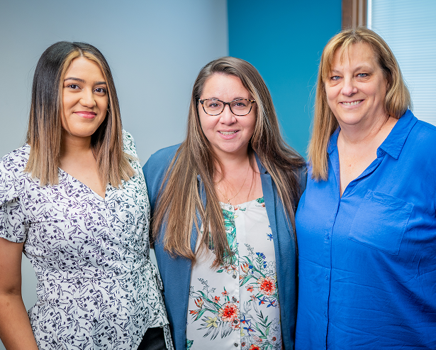 Three women stand indoors, smiling at the camera—capturing the welcoming spirit of Grand Junction Health Care. Each showcases her unique style, united by their shared commitment to providing affordable health care in the community.