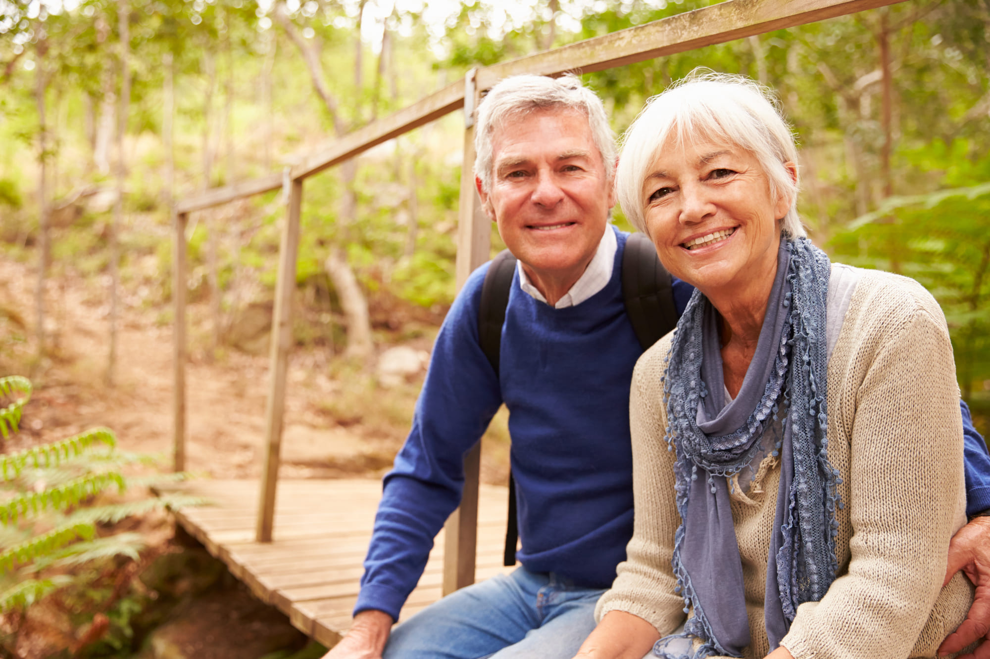 An older couple sits on a wooden bridge in a forest, smiling at the camera. Surrounded by lush greenery, they embody the happiness that comes with access to affordable health care in their golden years.
