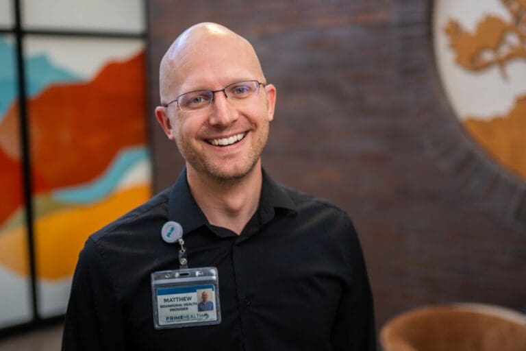 matthew bowen primehealth plus bald man with glasses and black shirt stands in front of colorful background