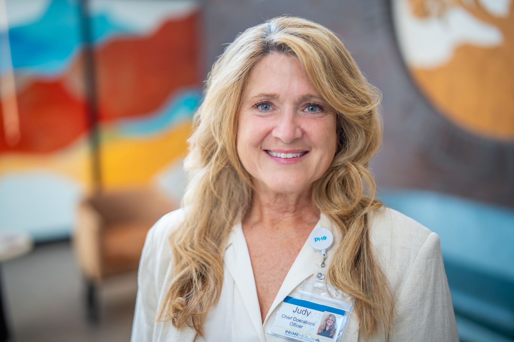 judy troyer chief operations officer for primehealth plus in grand junction, co woman with long blond hair and blue eyes looks at camera wearing cream jacket with blurred colorful background